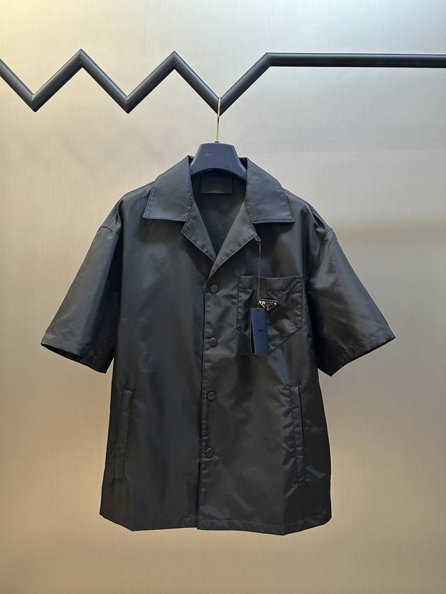 Prada 再生尼龙短袖衬衫 选用品牌标志性的再生尼龙面料，表面呈现丝绸光泽与细腻肌理，同时又保持相对轻盈的克重，与传统尼龙高强度，抗撕裂，耐磨损等特性相当的同