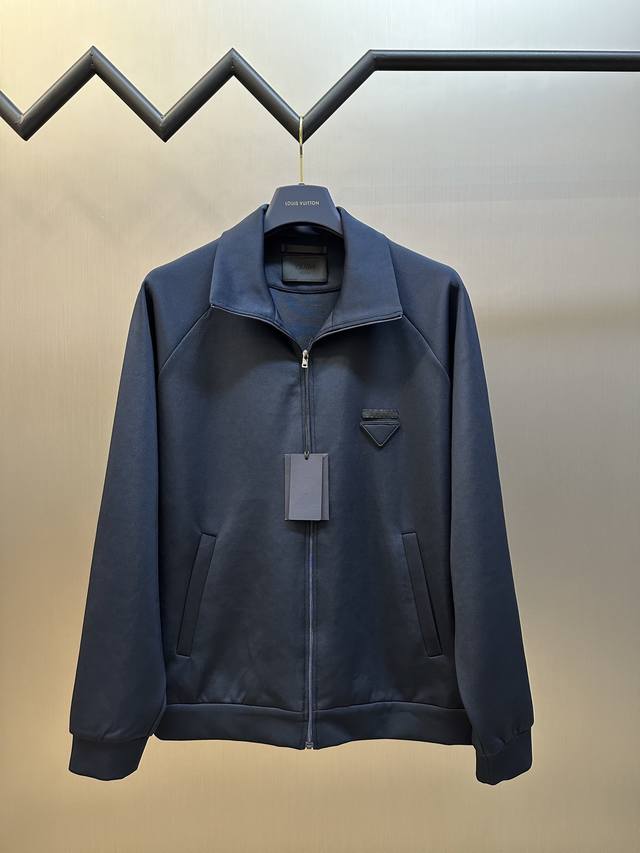 Prada 饰织物三角标夹克以80%天然棉纤维为主体，加入少量竹纤维与聚酯纤维等织造而成的健康布面料。棉纤维提供天然的透气性和舒适性，少量聚酯纤维和竹纤维的加入