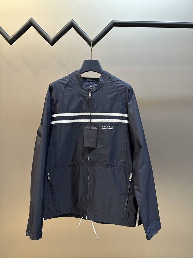 Prada 再生尼龙印花徽标夹克选用品牌标志性的再生尼龙面料，与传统尼龙性能相当，通过防污防水抗撕裂复合工艺处理之后，表面呈现独特的丝绸光泽触感，兼顾高强度，抗