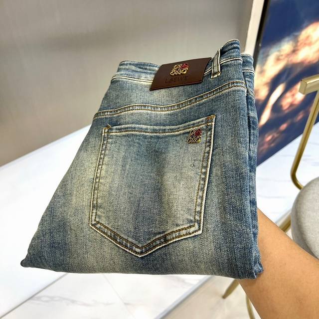 Lowe罗意威、2025秋冬新品牛仔裤，以独特的蓝灰色调呈现复古做旧质感。裤子采用精选牛仔面料，后袋饰有精致的金色金属品牌标识，腰间搭配棕色皮质标签，压印清晰品