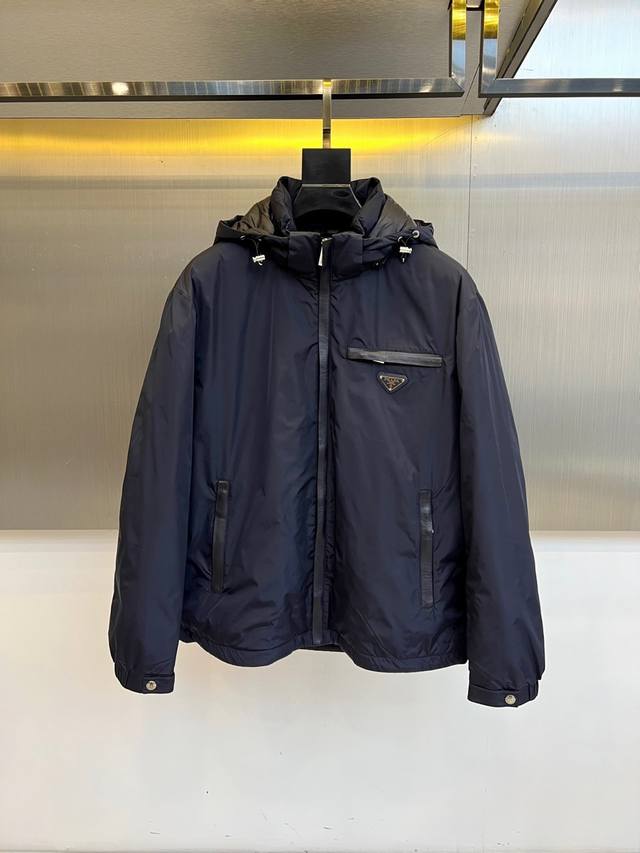 prada普拉、2025秋冬新品羽绒服，以经典黑色演绎现代极简美学。这款羽绒服采用100%聚酰胺面料打造，兼具出色保暖性能与轻盈质感。连帽设计搭配顺滑拉链开合系