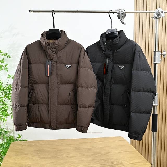 Prada普拉达2025冬季新款休闲百搭立领保暖面包羽绒服 这款羽绒外套采用创新的聚纤面料制作，颇具新颖特色。采用90%白鸭绒和10%羽毛填充具有较好的保暖性，