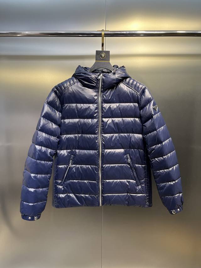 Prada连帽压纹垫肩测纹羽绒轻薄羽绒外套 这款羽绒外套采用现代廓形，融合了运动风格与时尚细节。休闲设计点缀涂珐琅三角形金属徽标，搭配技巧连帽和90绒填充物填充