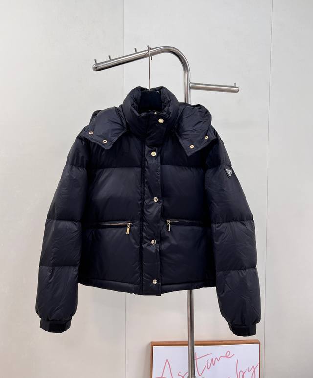 prad25Fw秋冬三角标连帽羽绒服外套，抽绳末端的金属扣为品牌定制款，质感细腻且经久耐用。门襟选用双向金属拉链，齿牙精密咬合，拉合顺畅度经过多轮测试。衣身填充