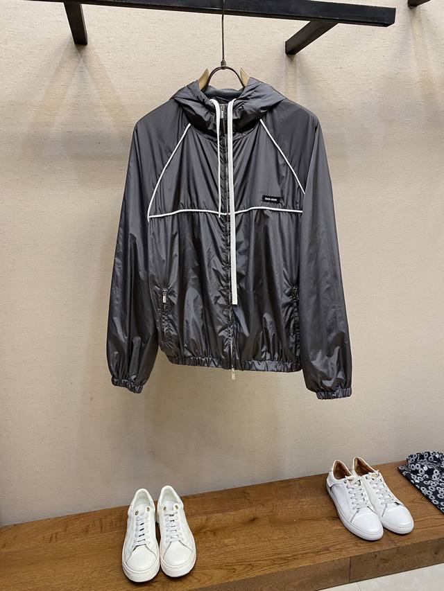 Miumiu 拉链风帽夹克 定制冷萃智孔纤维尼龙精纺布 轻薄细腻 舒适贴肤 以经典黑为主色调 深達与高贵的极致融合 醒目的Miumiu徽标贴饰点缀于胸前 展现出